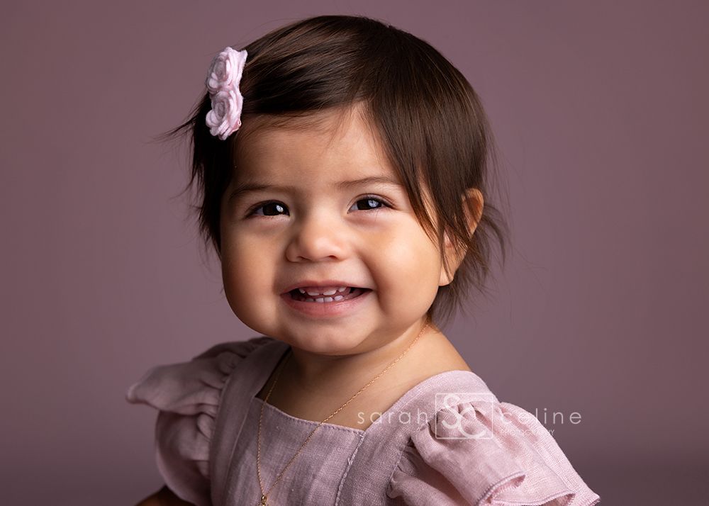 A child in a pink dress smiles and plays during a professional portrait photography session against a mauve backdrop.