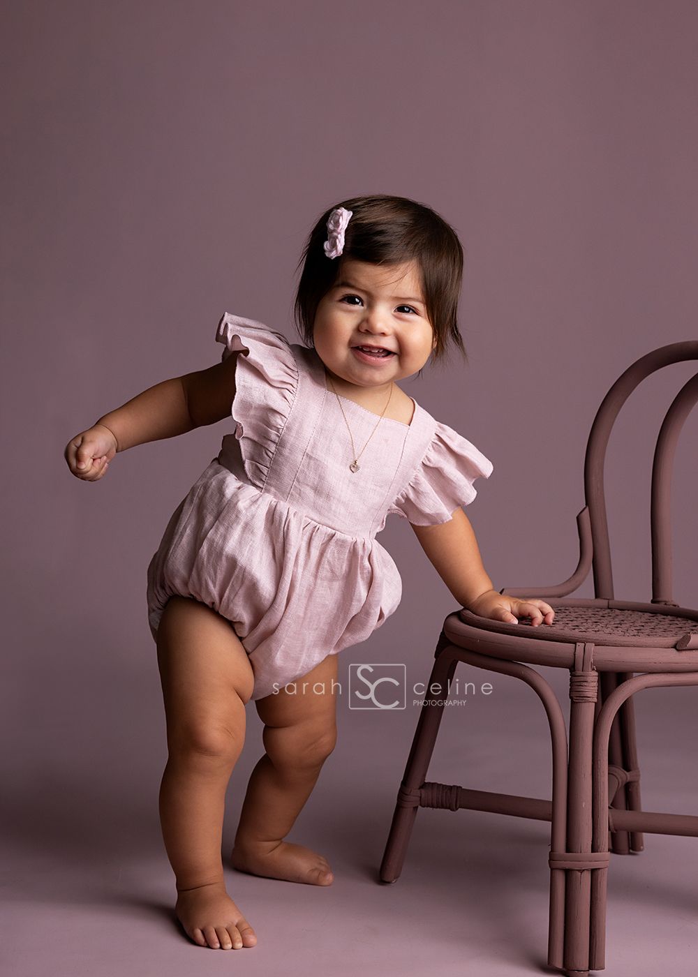 A small child in a pink ruffled romper stands playfully next to a brown rattan chair against a mauve backdrop.