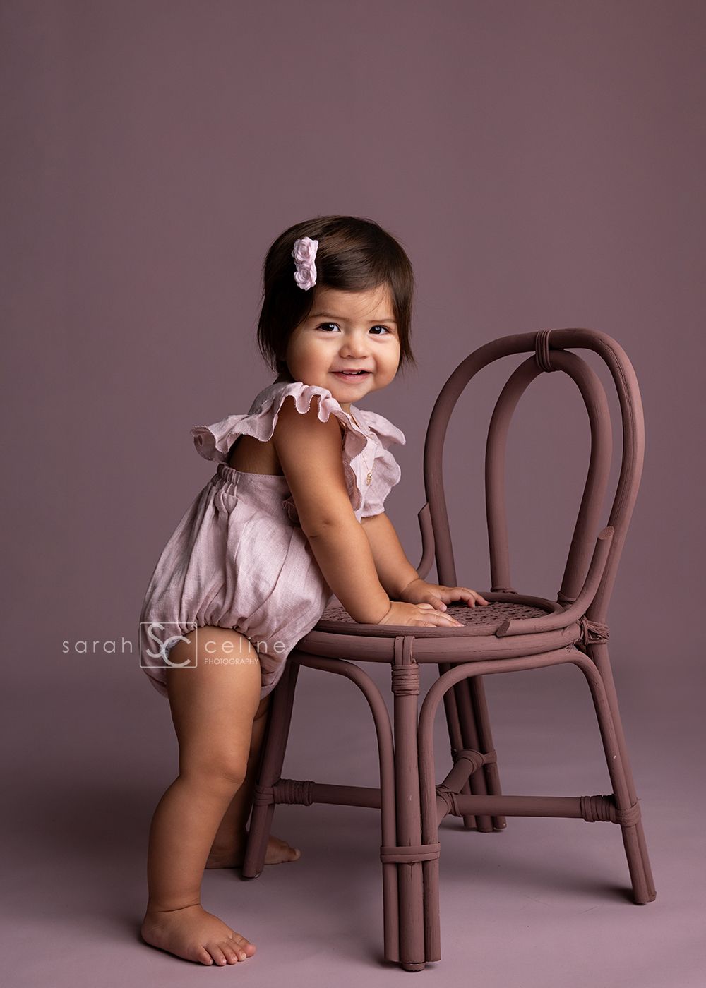 A toddler in a pink romper leans against a brown rattan chair during a studio photoshoot with mauve background.