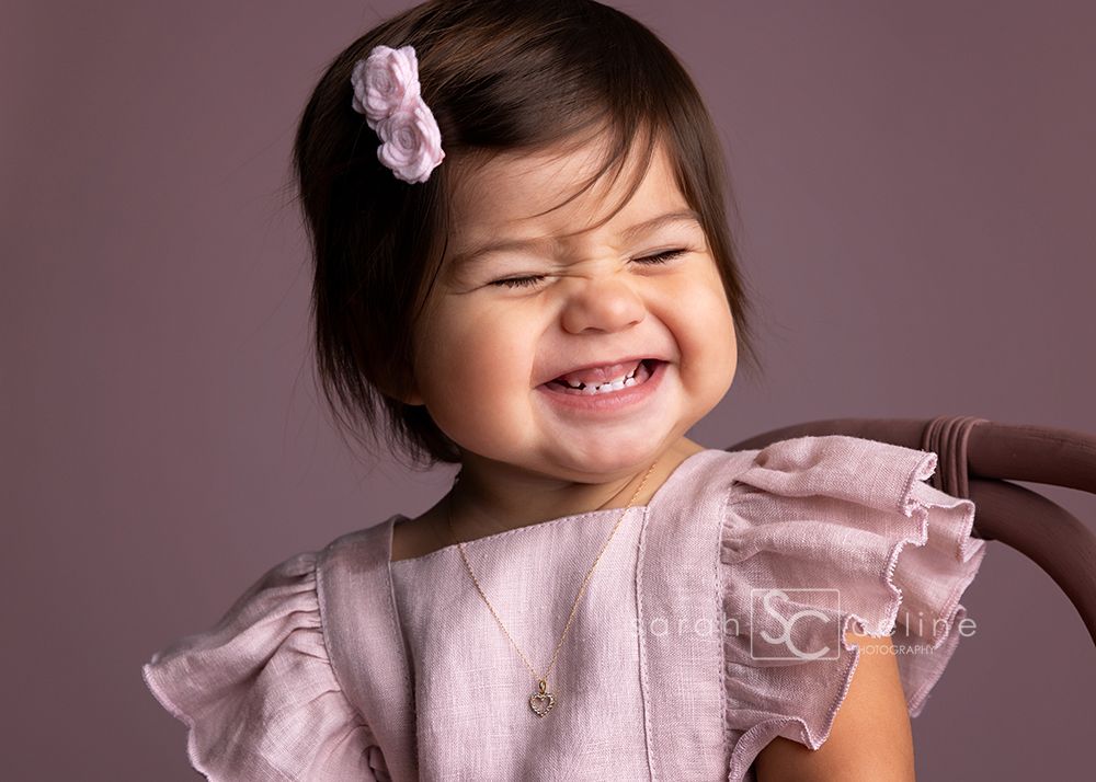 A joyful expression captured in a close-up portrait wearing a pink ruffled outfit against a mauve background.