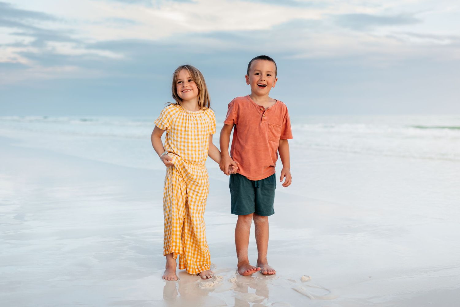 Two figures walking hand in hand on pristine beach with gentle waves and cloudy sky.