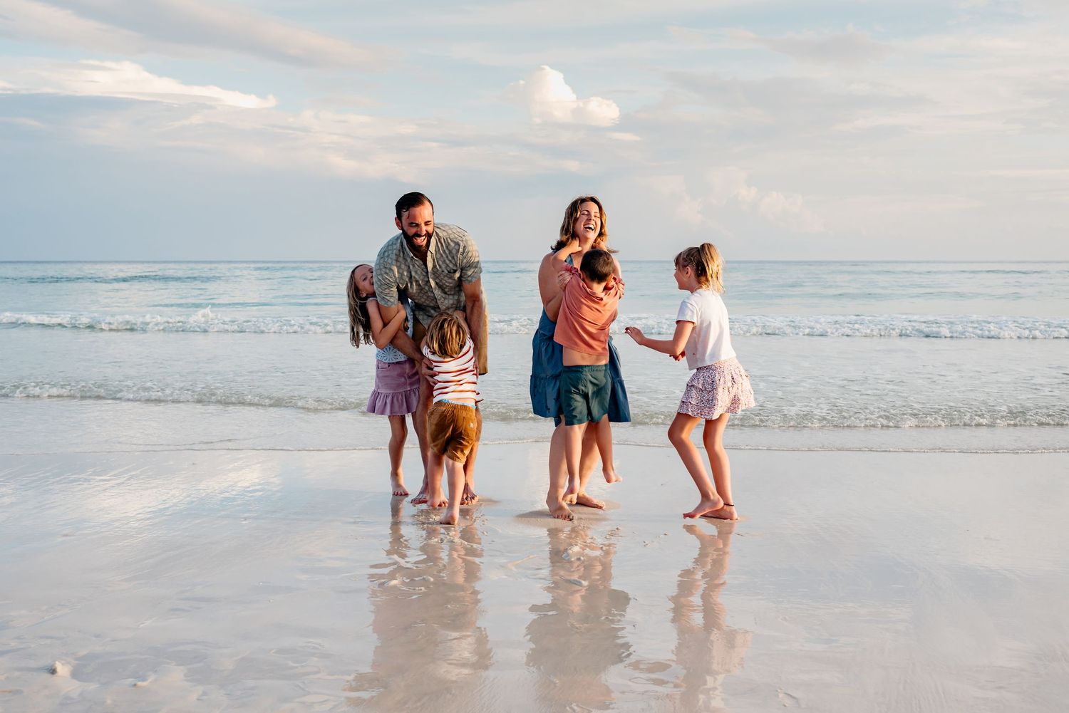 Family enjoying a sunset walk along the shoreline of a beach with gentle waves and soft clouds in the background.