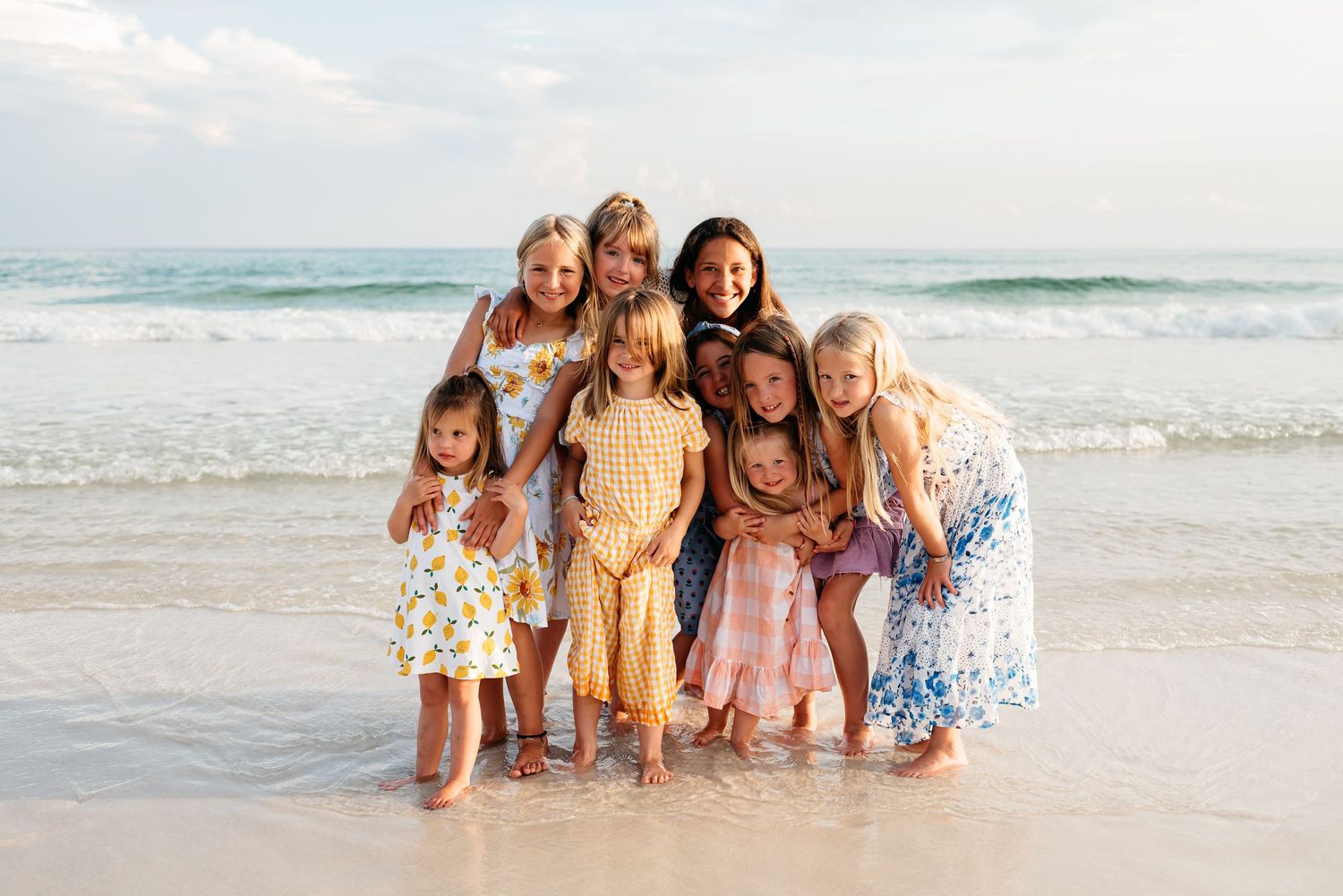 A group of people wearing coordinated yellow and pink outfits pose together on a sunny beach by the ocean waves.
