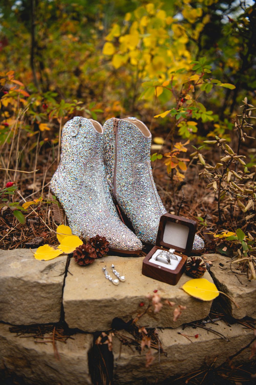 Sparkly silver boots rest on a stone ledge next to a ring box in an autumn outdoor setting with yellow leaves.