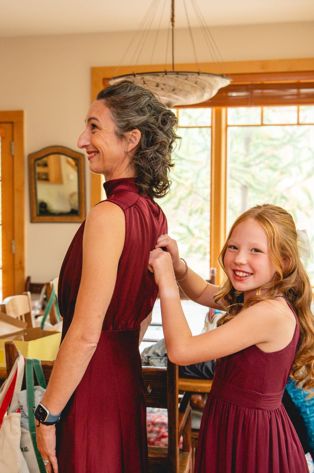 People wearing burgundy dresses share joyful moments while getting ready in a sun-lit room with wooden trim windows.