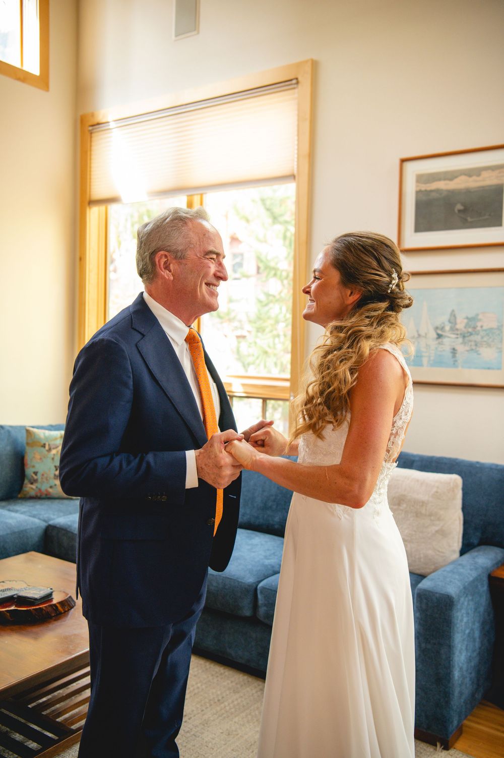 In a bright room, two figures share a tender moment near a blue sofa and window with one wearing a navy suit and white dress.