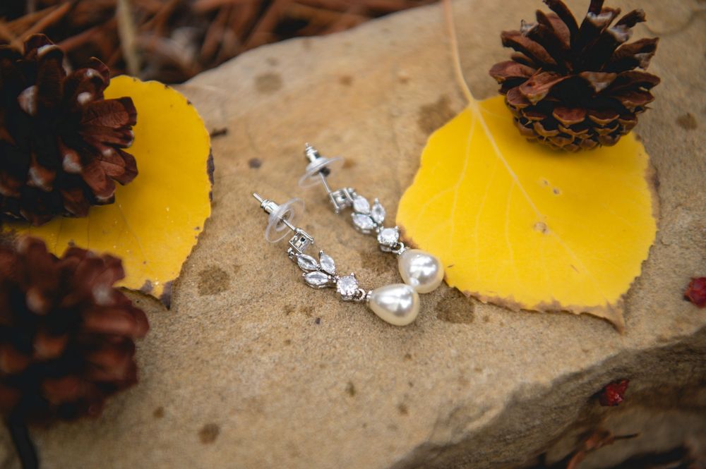 Crystal earrings sparkle among yellow autumn leaves and pine cones on natural stone surface.