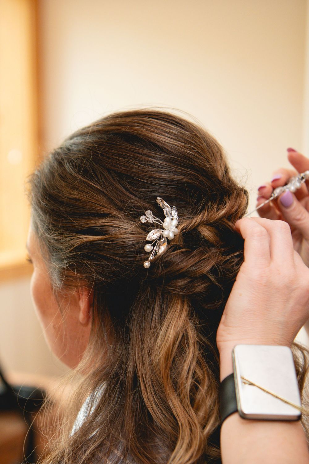A wedding hairstyle being styled with delicate crystal hair accessories and braided details in a warm-lit room.