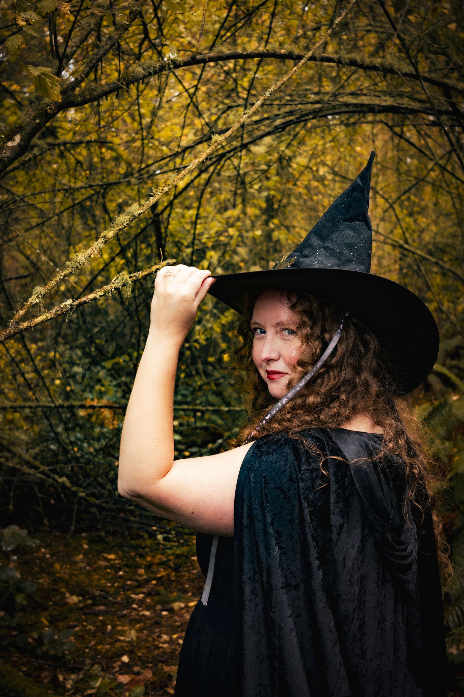 Woman wearing witch hat and costume in the forest looking at the camera.