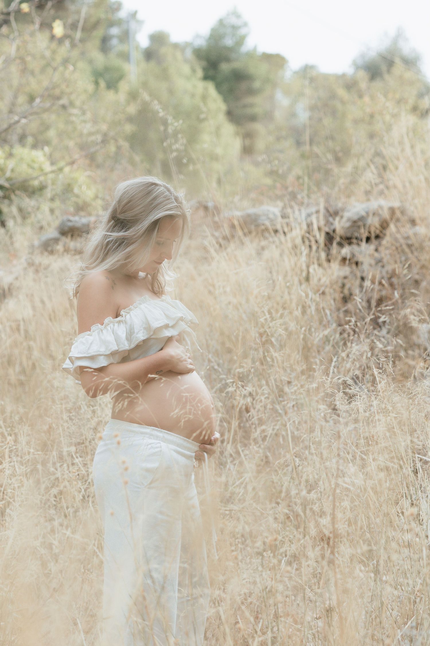 Ethereal maternity portrait in a sun-drenched field of tall golden grass wearing delicate white clothing.