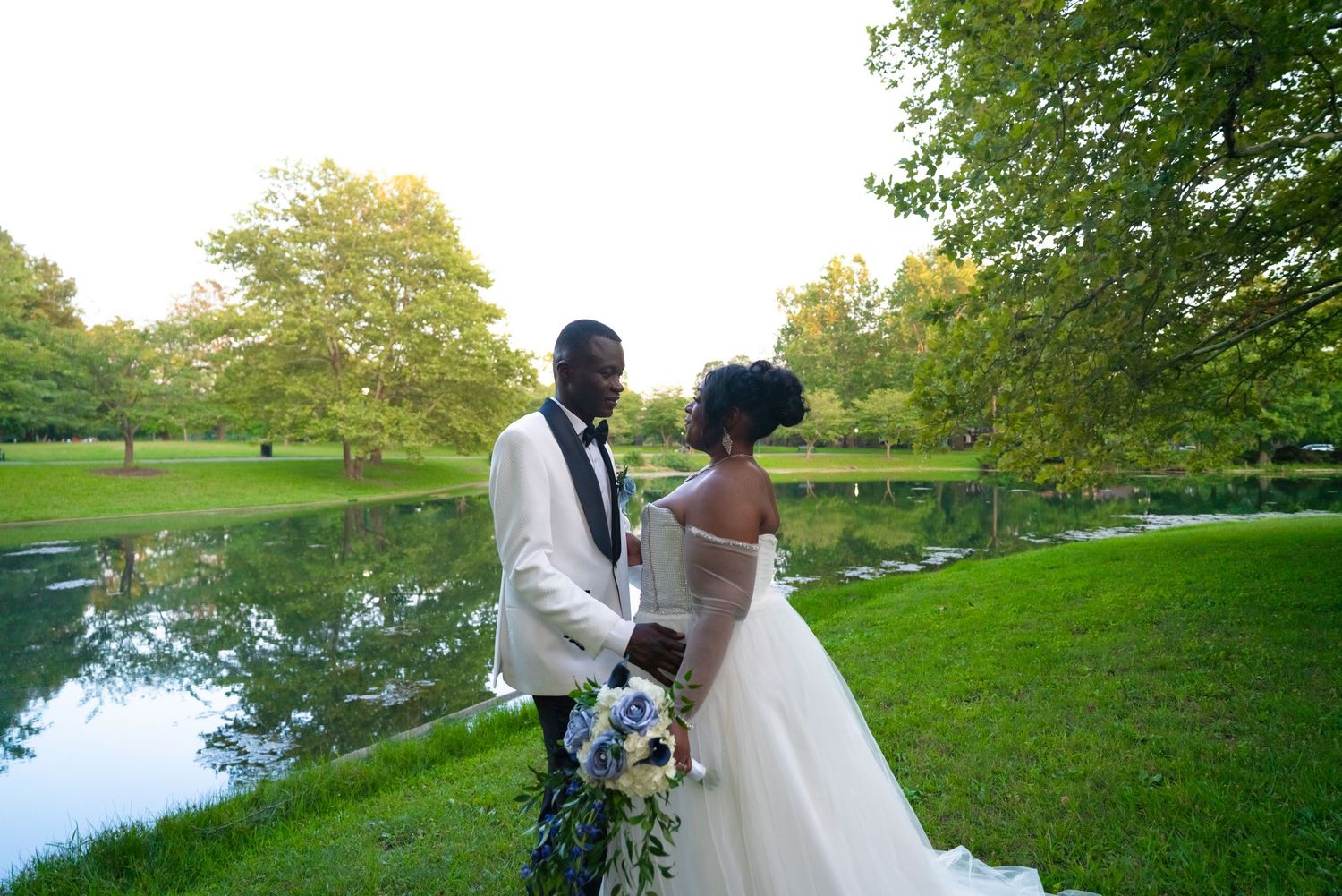 A bride and groom pose together by a serene pond surrounded by lush greenery.