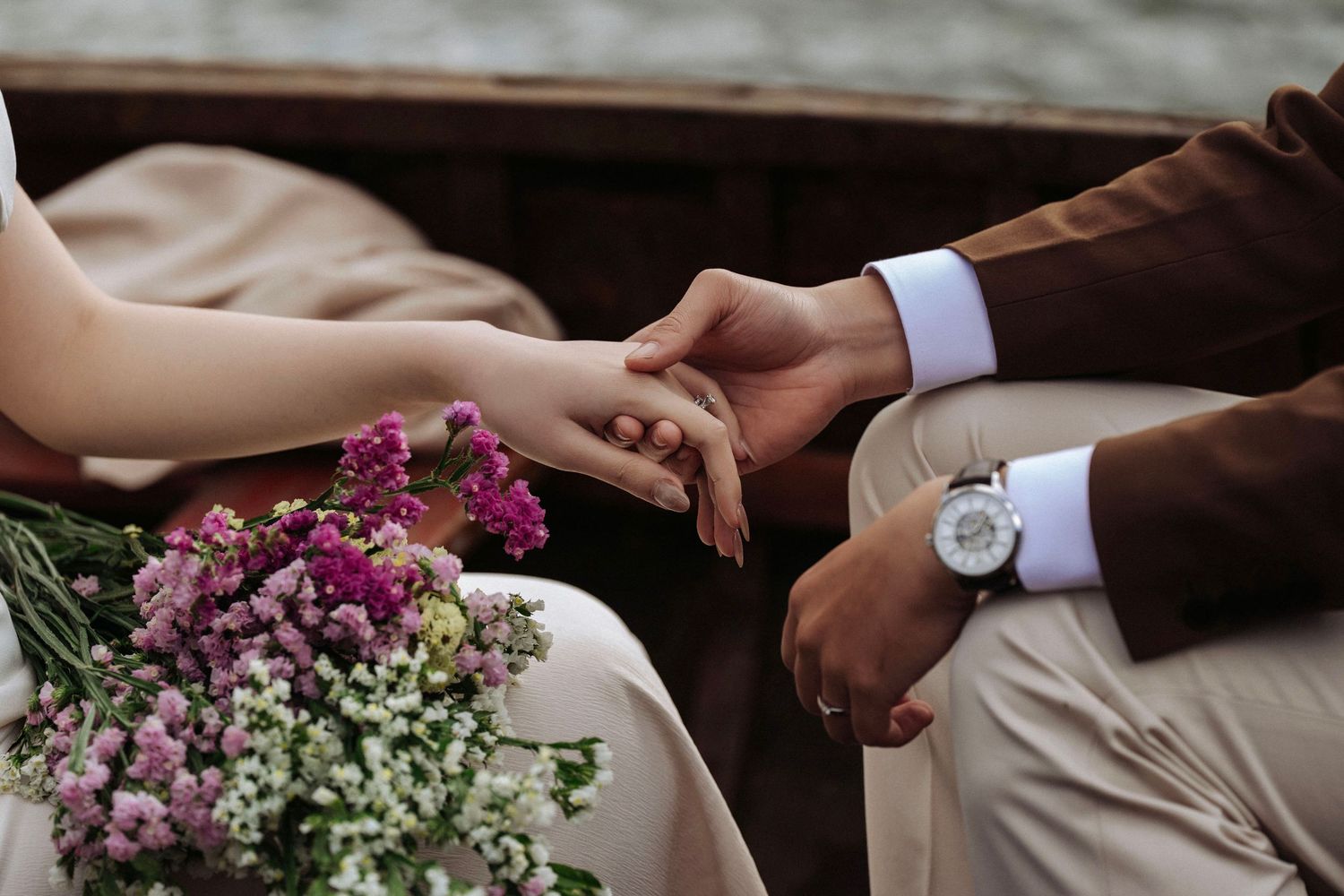 Close-up of couple holding hands on a boat with pink and white flowers, groom wearing a watch and wedding ring.