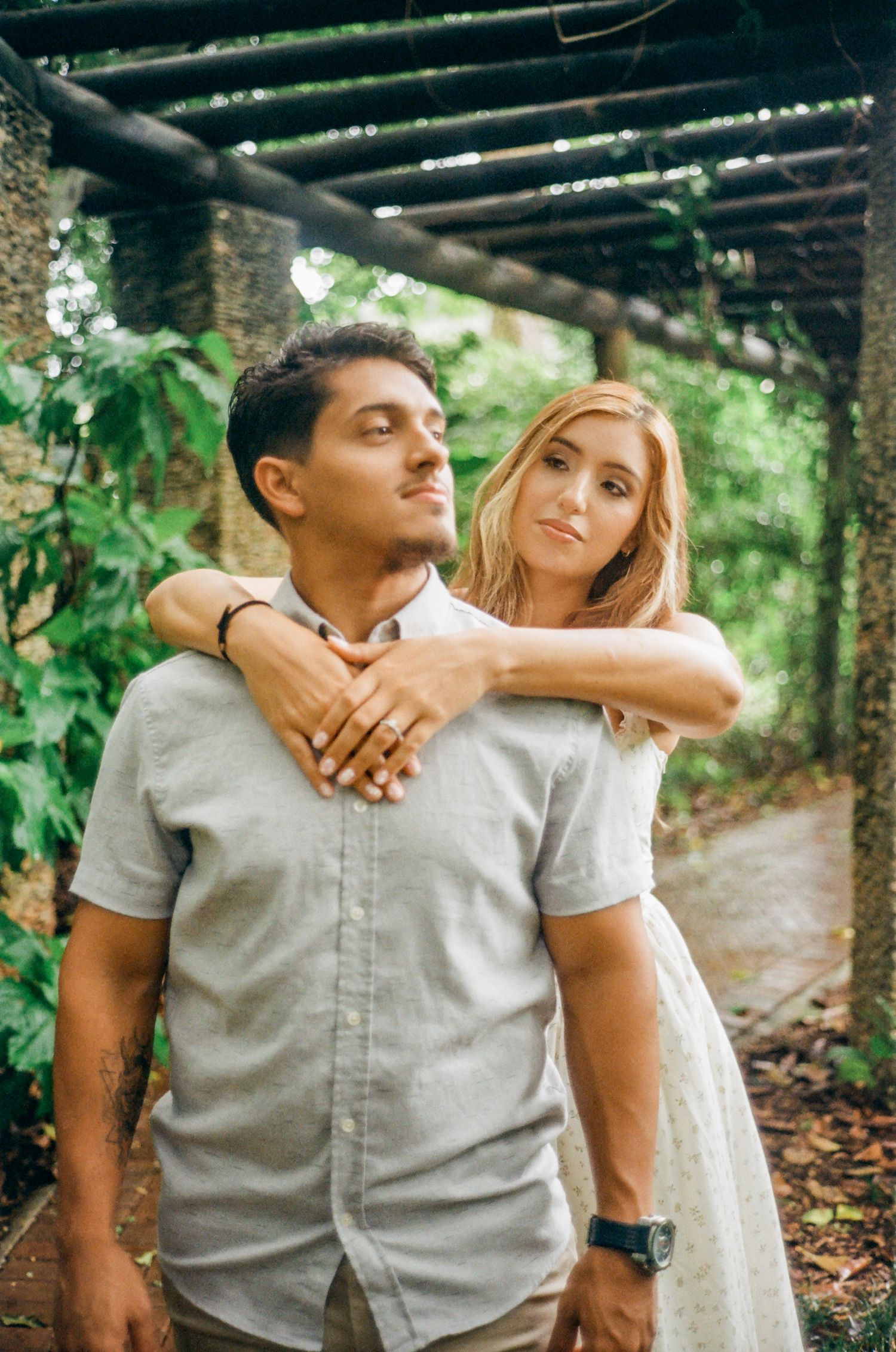 A romantic couple embraces under a rustic wooden pergola surrounded by lush greenery.