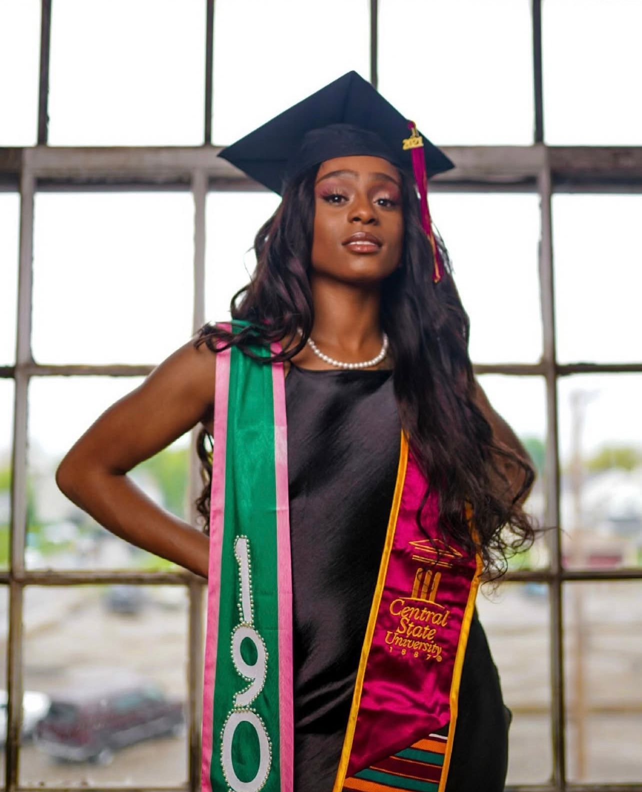 Graduate wearing graduation cap and stoles poses confidently by industrial windows.