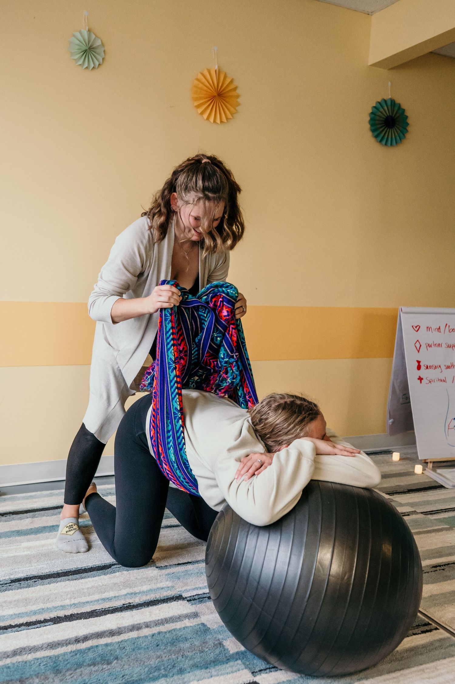 Doula using a rebozo scarf technique on pregnant woman leaning over birth ball during labor support session.
