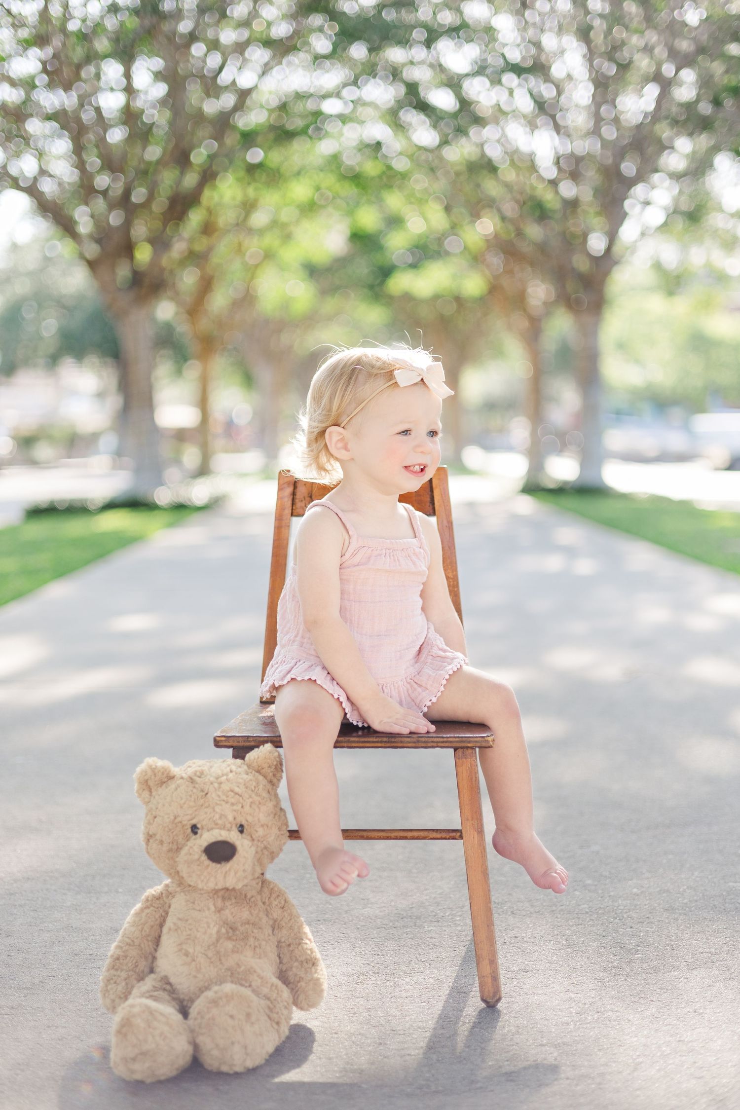 A toddler in a pink romper sits on a wooden chair next to a teddy bear on a tree-lined path.
