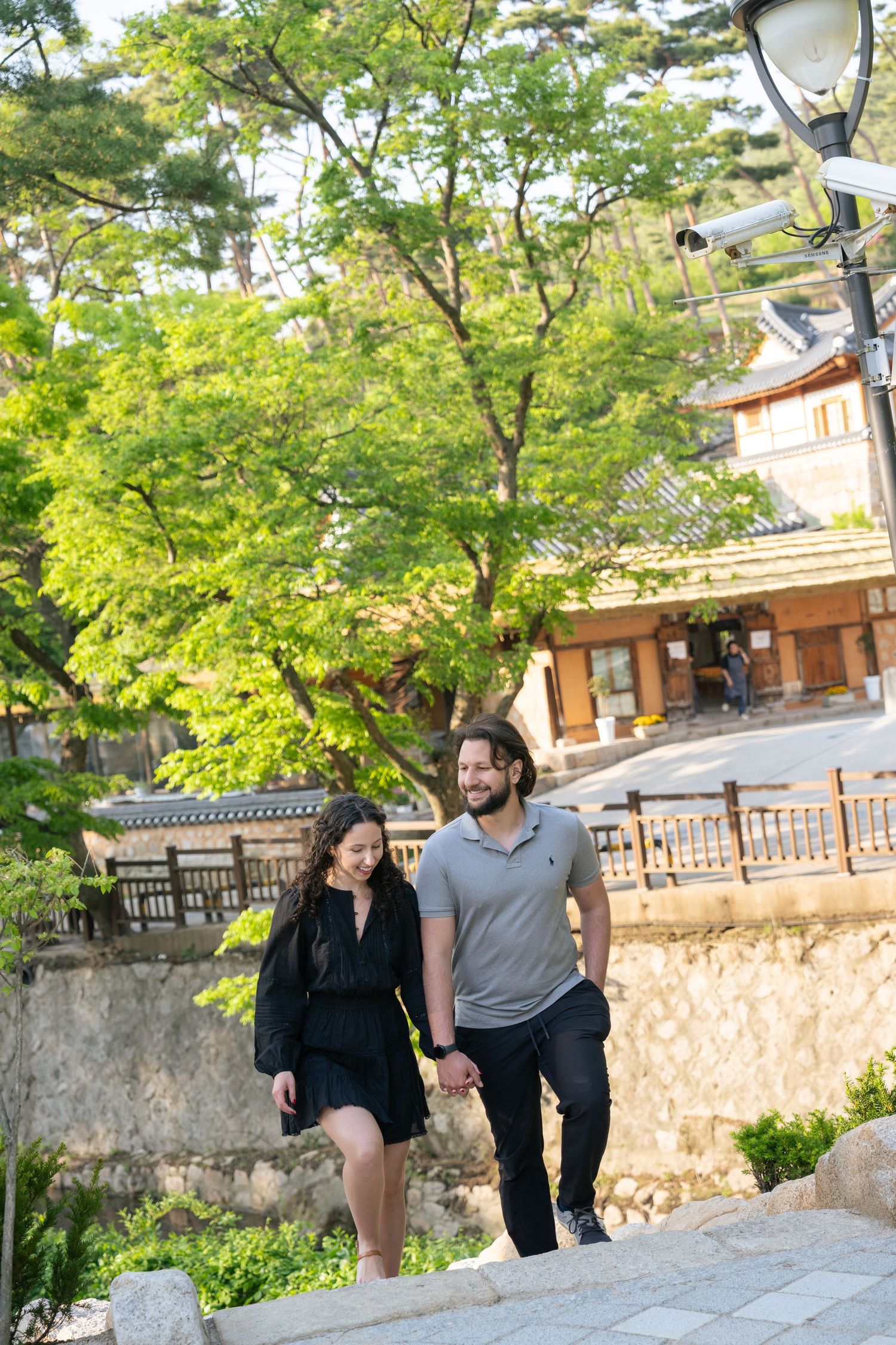 A couple takes a romantic walk along a scenic stone path lined with lush green trees in a peaceful garden setting.