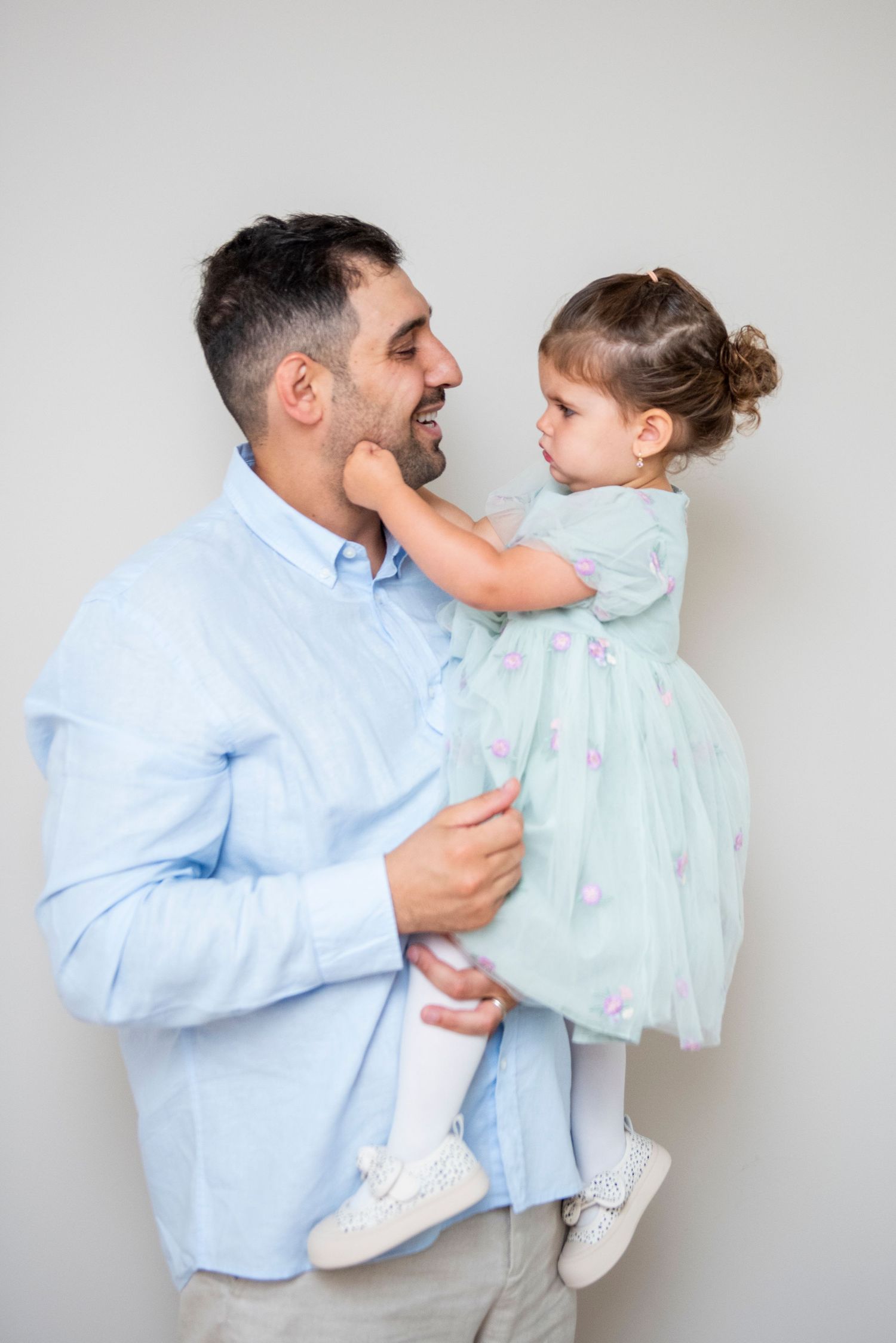 Father in light blue shirt playfully interacting with toddler in mint green dress.