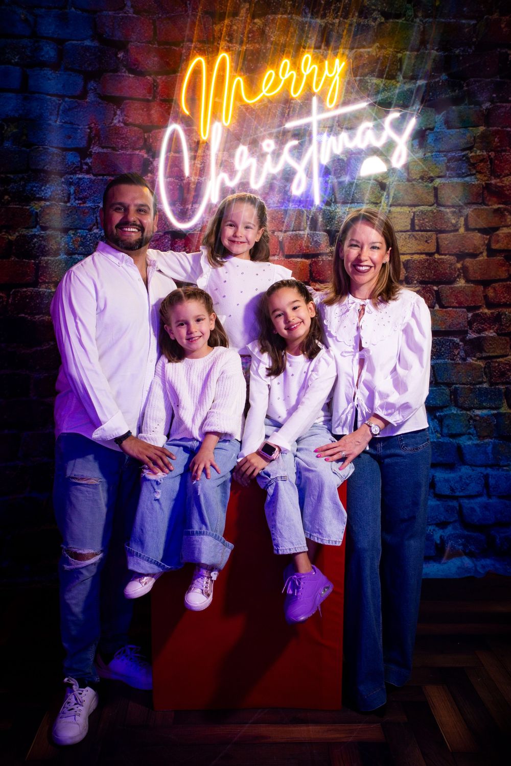 A family wearing matching white shirts poses together against a brick wall with neon lighting.