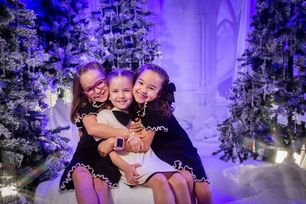 Three people pose together for a holiday photo surrounded by snow-covered Christmas trees and purple lighting.