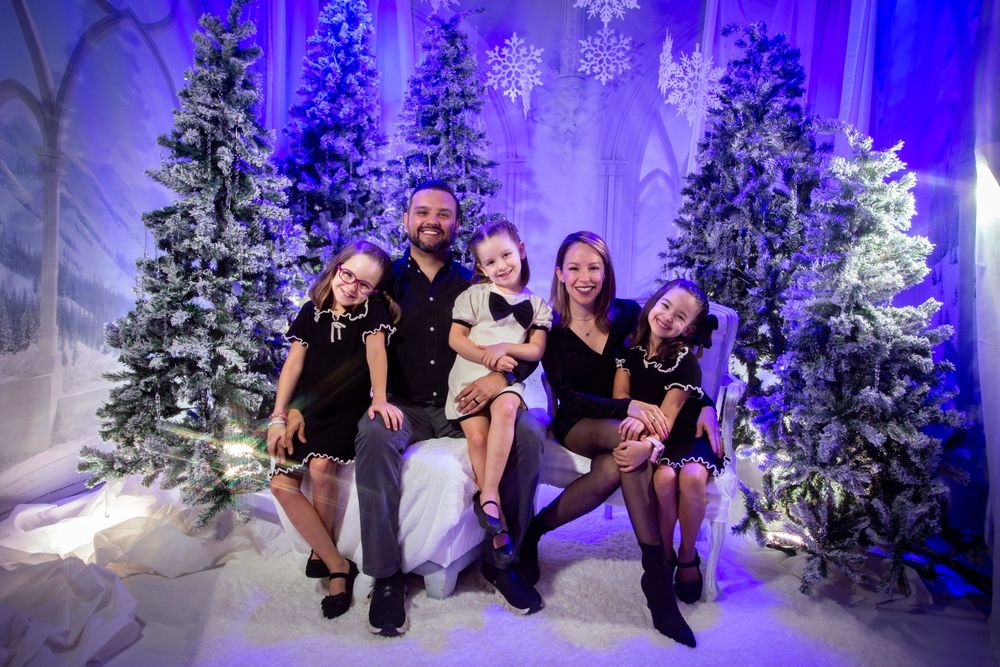 Group poses with wide smiles in front of snow-covered Christmas trees illuminated by blue lighting.