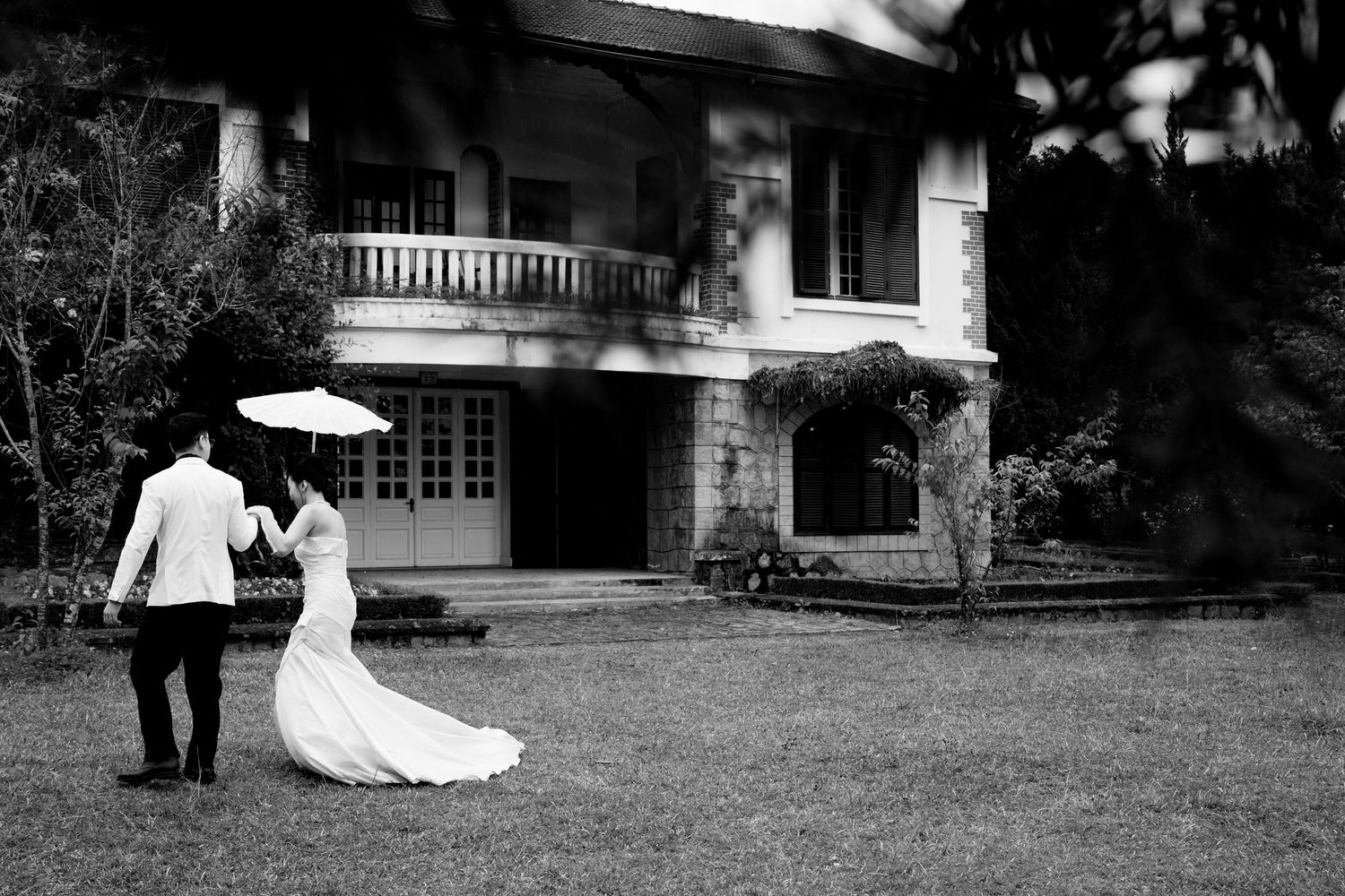 Bride and groom walk hand in hand across a lush lawn toward an elegant colonial mansion.