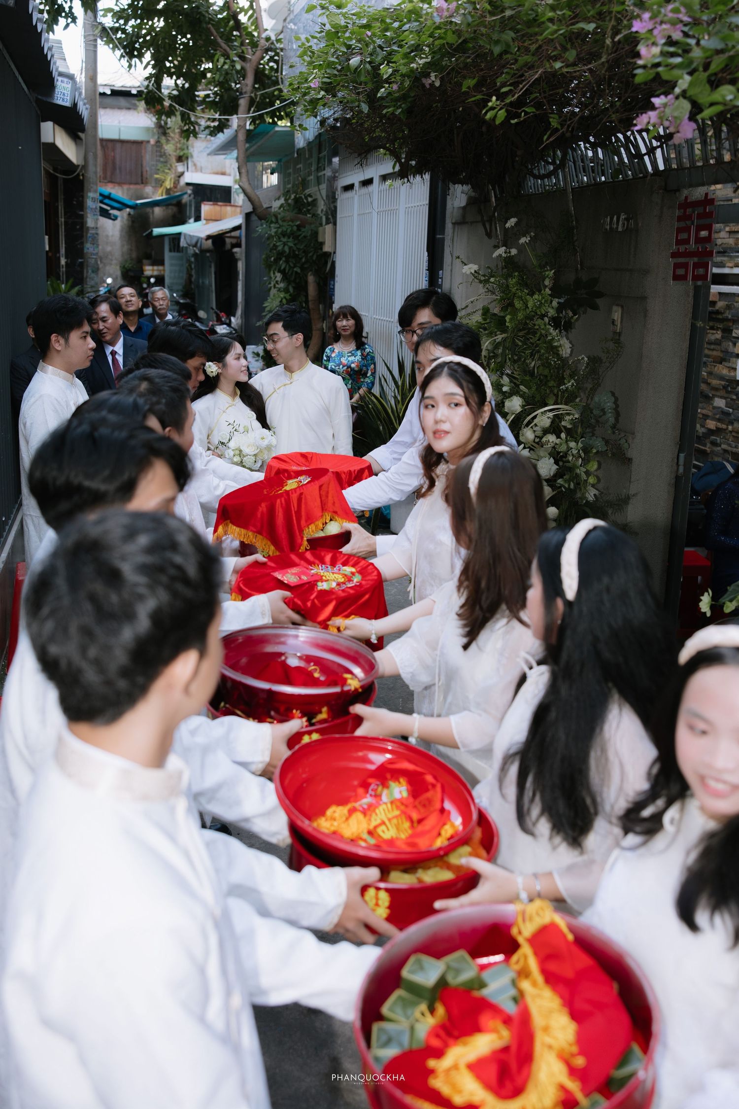 Vietnamese bride and wedding party in white ao dai passing red lacquer gift trays in an alley.