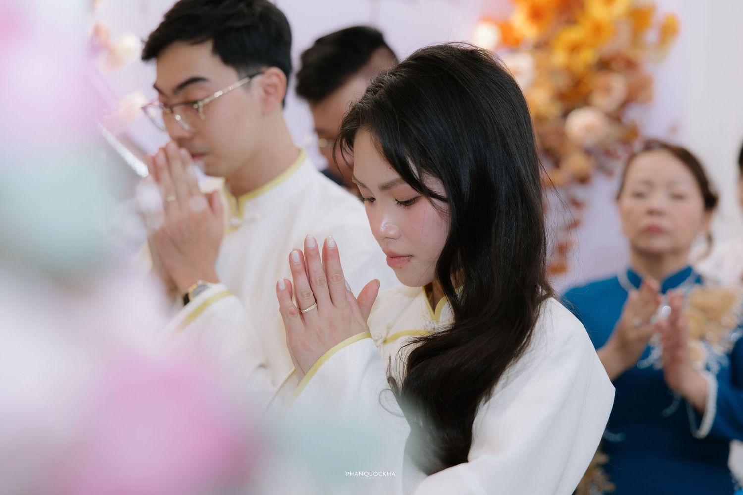 Vietnamese bride and groom in white ao dai pray with hands together at wedding ceremony.
