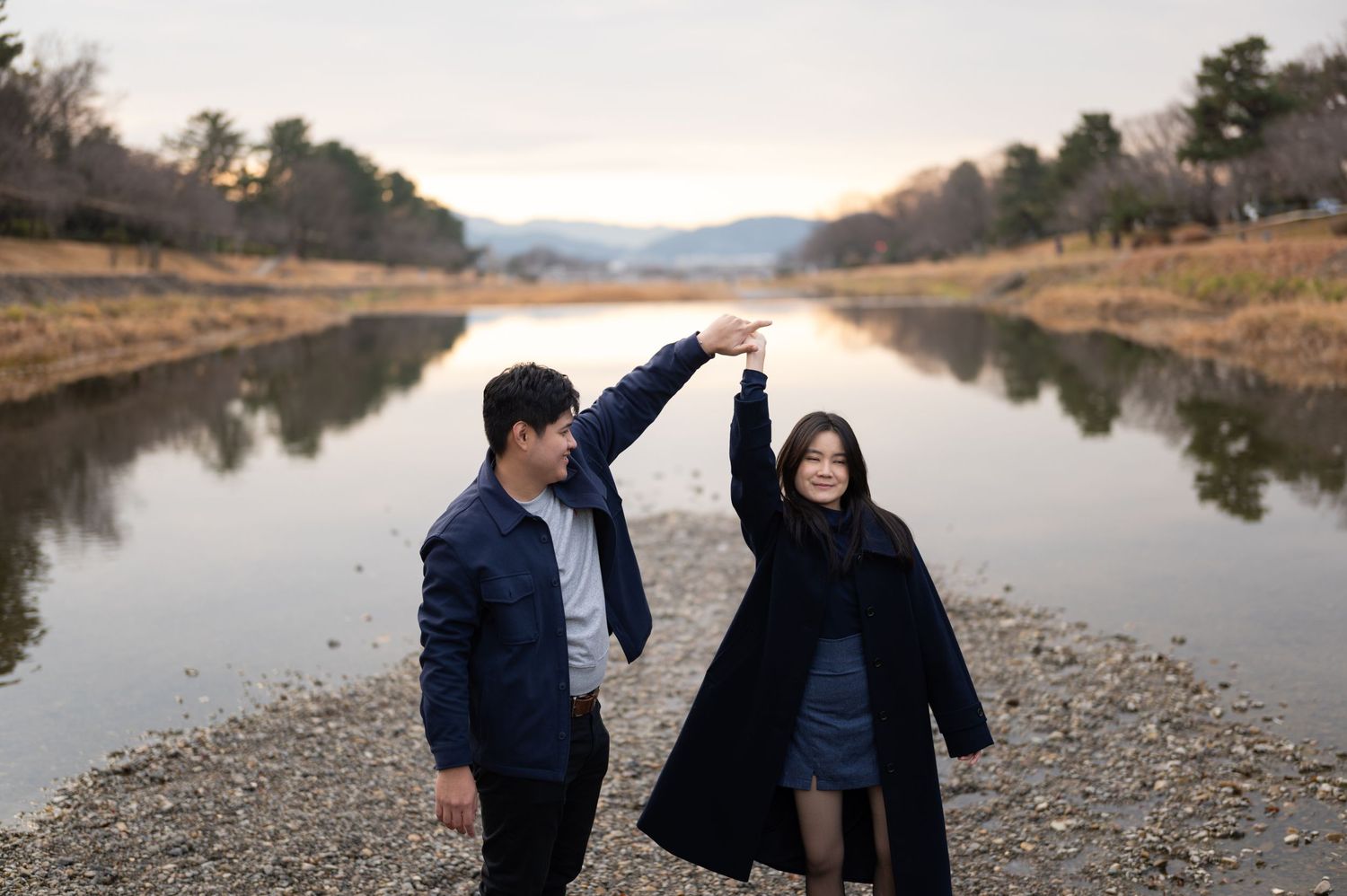 Couple posing playfully by a tranquil river, man raising woman's hand, with mountains behind.