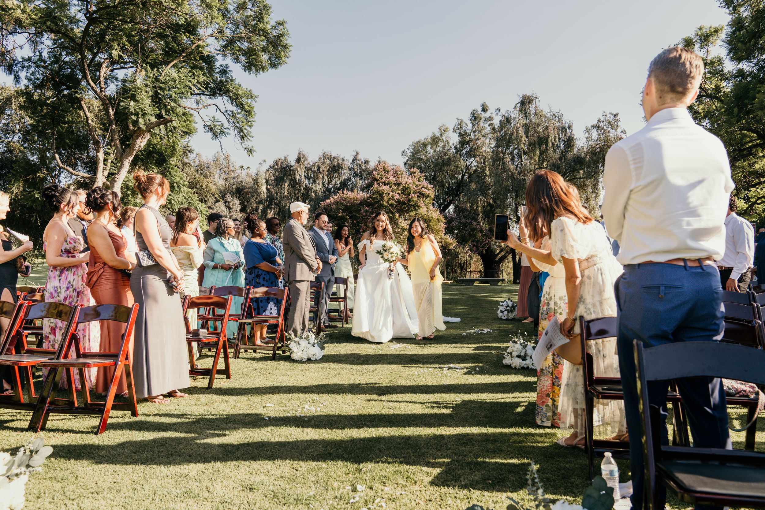 Guests seated outdoors at rustic wooden tables during a sunny garden wedding reception.