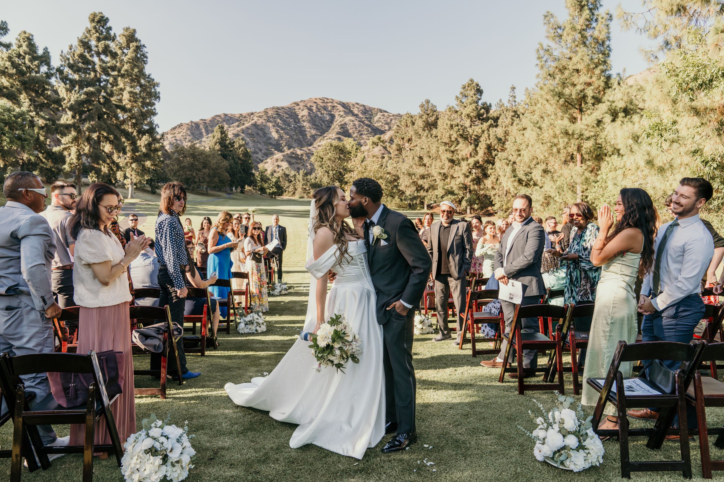 Outdoor wedding ceremony in a mountain setting with guests seated on chairs and floral decorations.