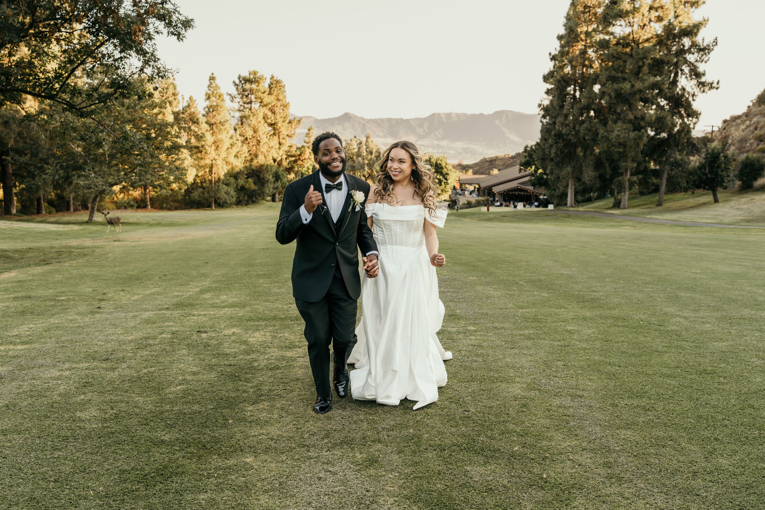 Wedding couple walks hand in hand across a sunlit golf course against mountain backdrop.