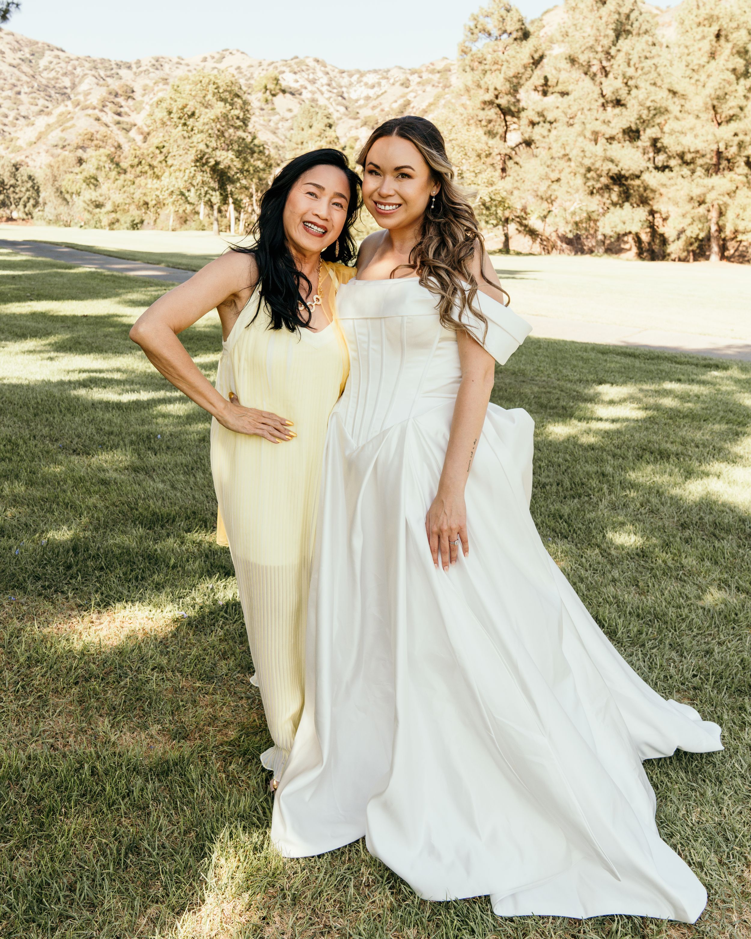 Two women in yellow and white dresses stand together on grass with trees and mountains in the background.