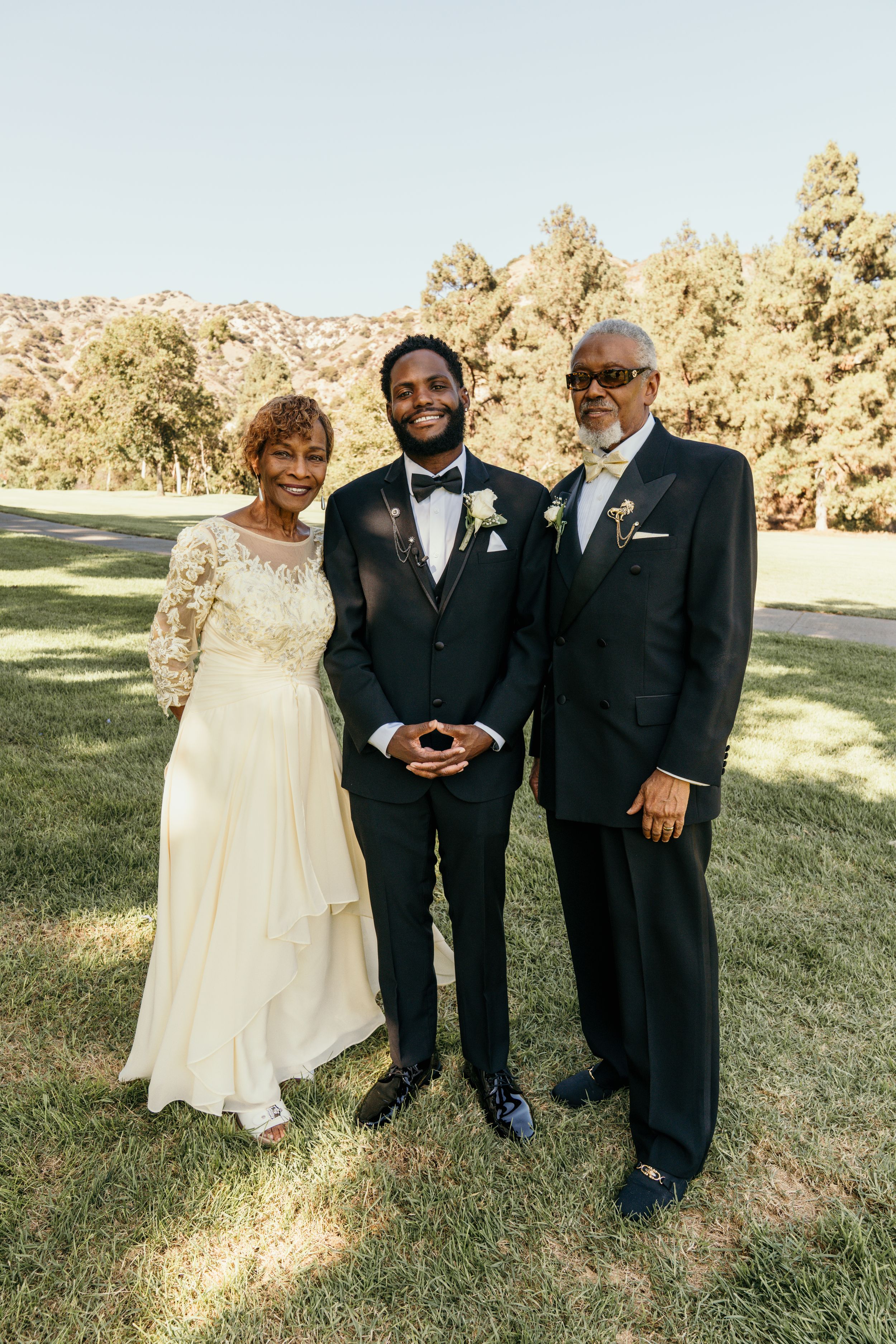 Three wedding guests in formal attire pose together on a sunny lawn during an outdoor celebration.