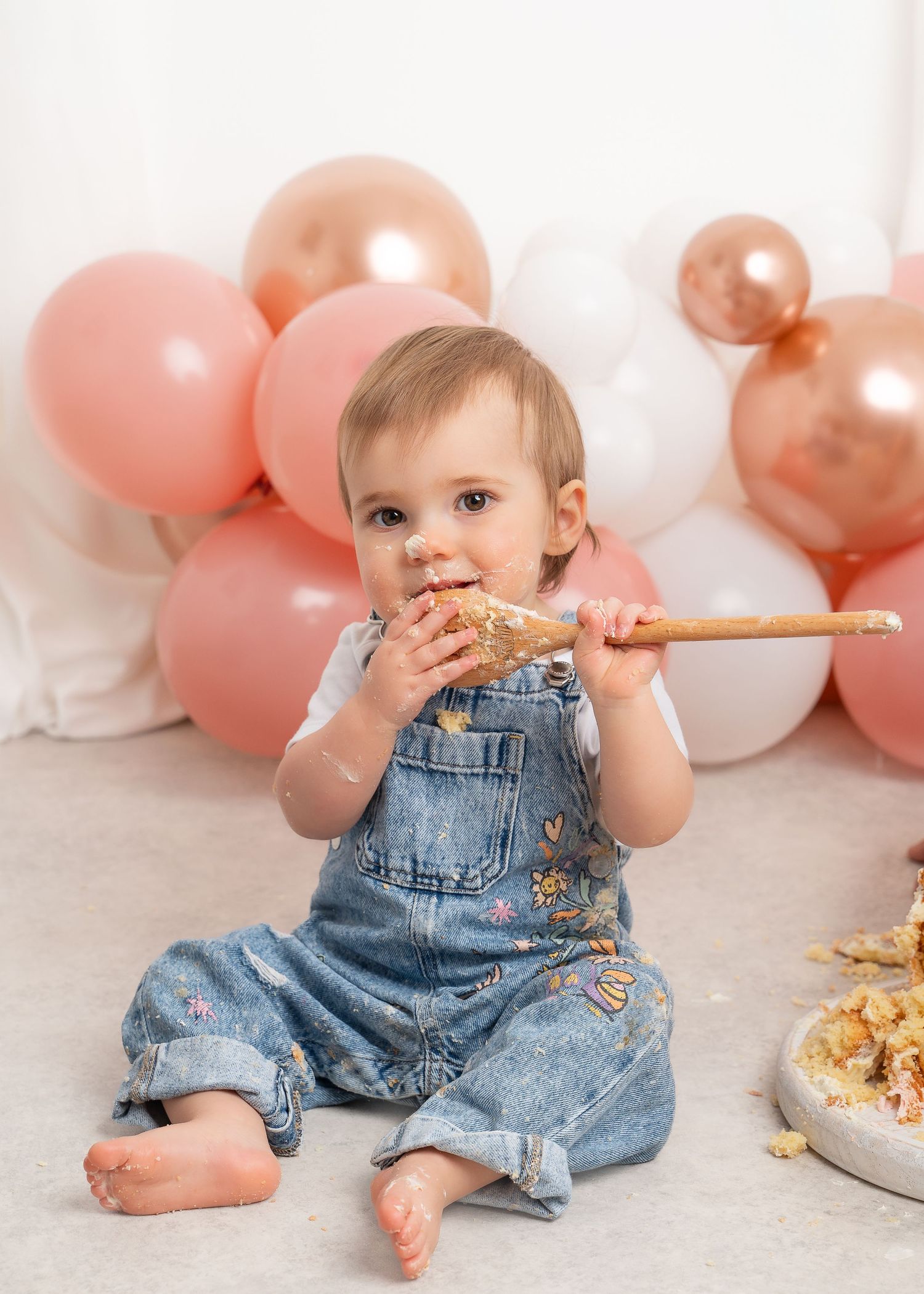 Baby in denim overalls plays with cake during cake smash photo shoot with pink balloons in Lanarkshire.