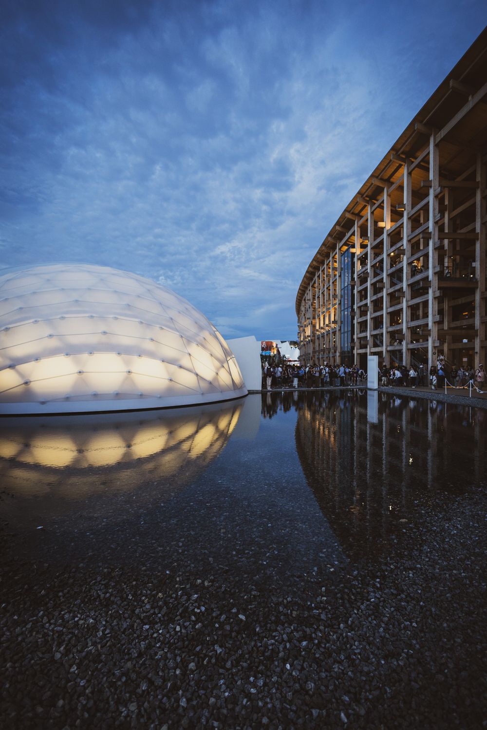 Glowing dome pavilion reflected in wet pavement beside a modern wooden structure at twilight.
