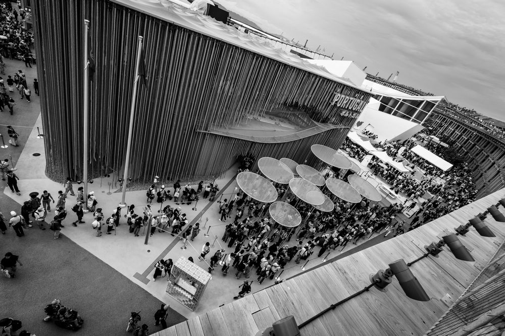 Aerial black and white view of outdoor cafe with umbrellas and crowds at a large venue.