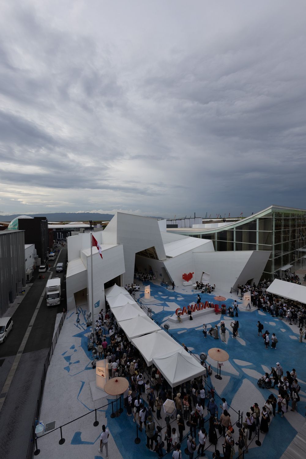 Aerial view of a white angular modern pavilion surrounded by crowds under cloudy skies.