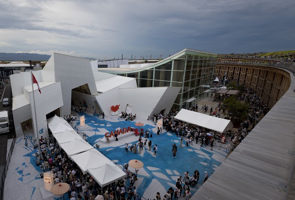 Overhead view of a white geometric pavilion with crowds and tents under overcast skies.