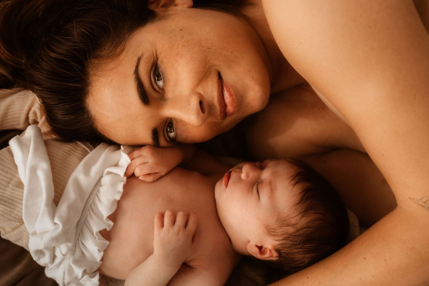 A tender moment between parent and newborn in Javea, baby lying together on white bedding in warm lighting- Javea newborn photographer