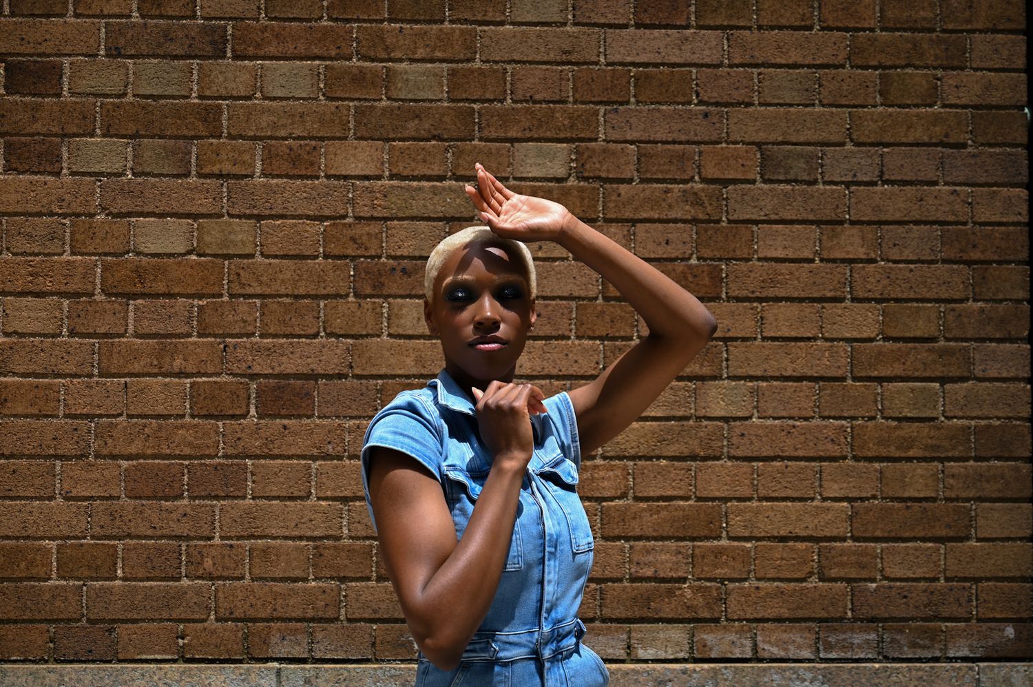 Person in a blue denim top poses expressively against a brick wall background.
