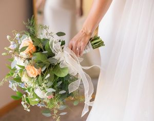 A delicate bridal bouquet featuring white and peach flowers with lush greenery and flowing ribbon accents. Columbus weddings.