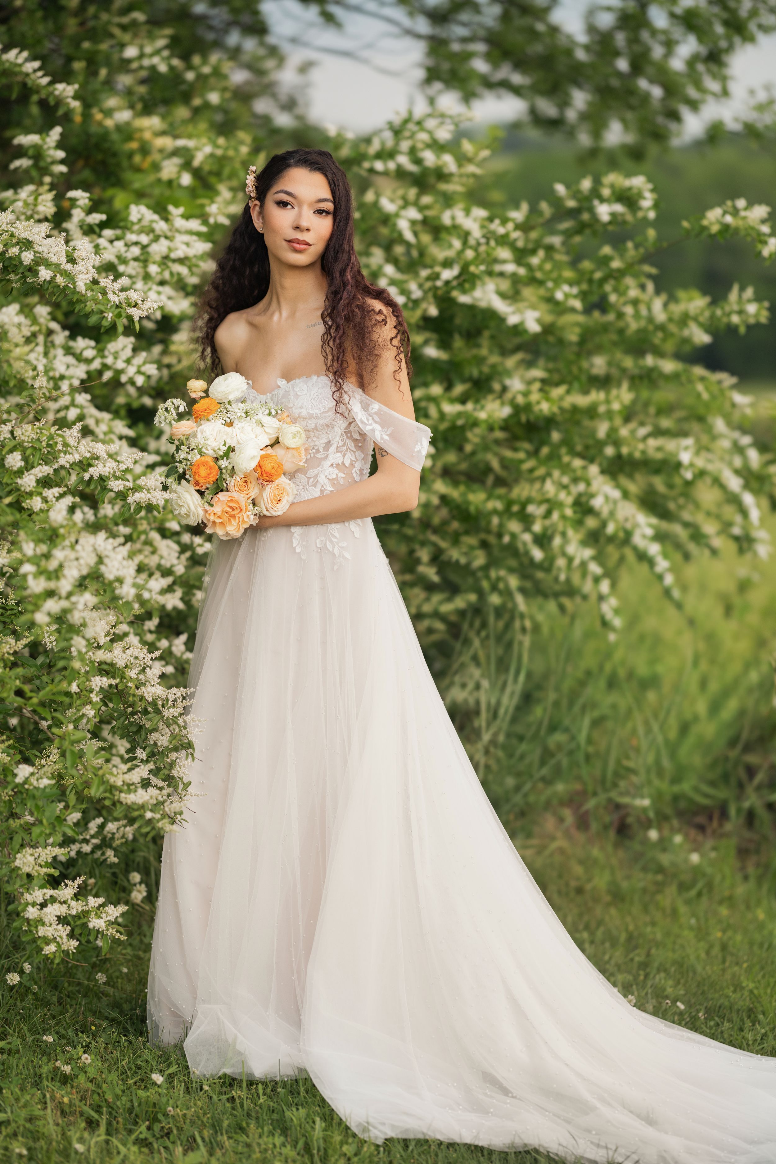 A bride in a flowing white tulle gown poses gracefully among blooming spring dogwood trees with a peach and white bouquet. Columbus, Ohio.