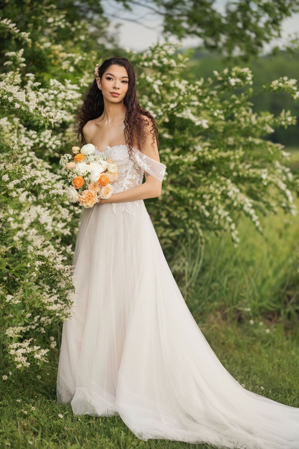A bride in a flowing white tulle gown poses gracefully among blooming spring dogwood trees with a peach and white bouquet. Columbus, Ohio.