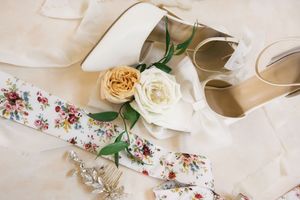 Elegant bridal accessories including white and peach roses, floral garter, and ivory wedding shoes arranged on light fabric.
