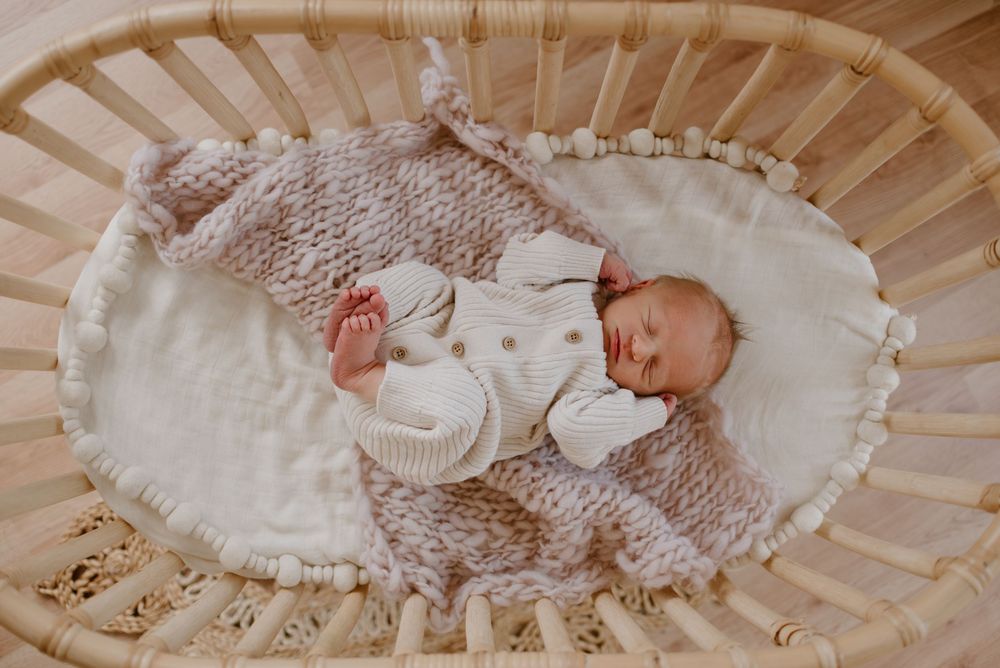 Newborn baby sleeping peacefully in white knit outfit on cream-colored bassinet with ruffled trim.