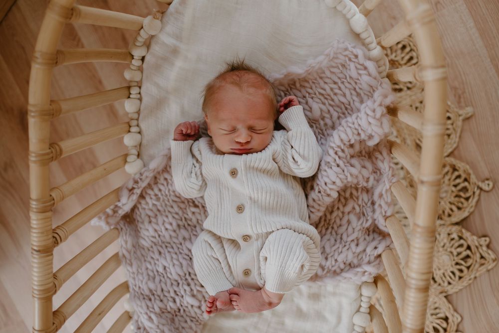 Sleeping newborn wearing white cardigan rests in wooden bassinet with knitted blanket.