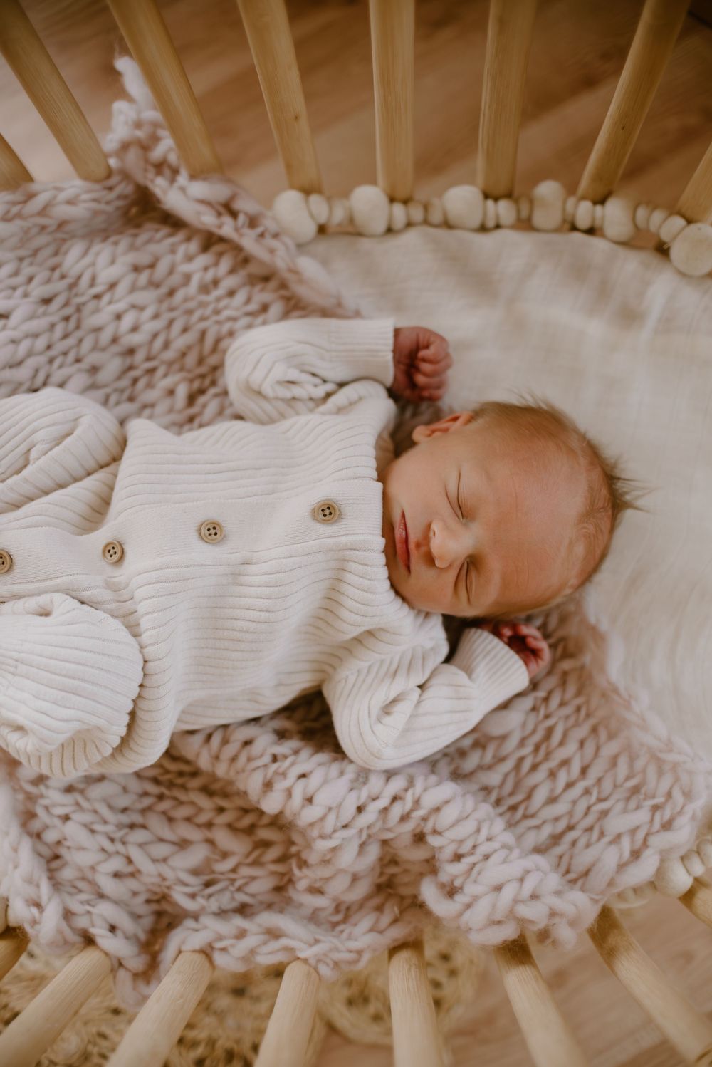 Peaceful newborn in white knitted outfit sleeping in wooden bassinet with textured blanket.