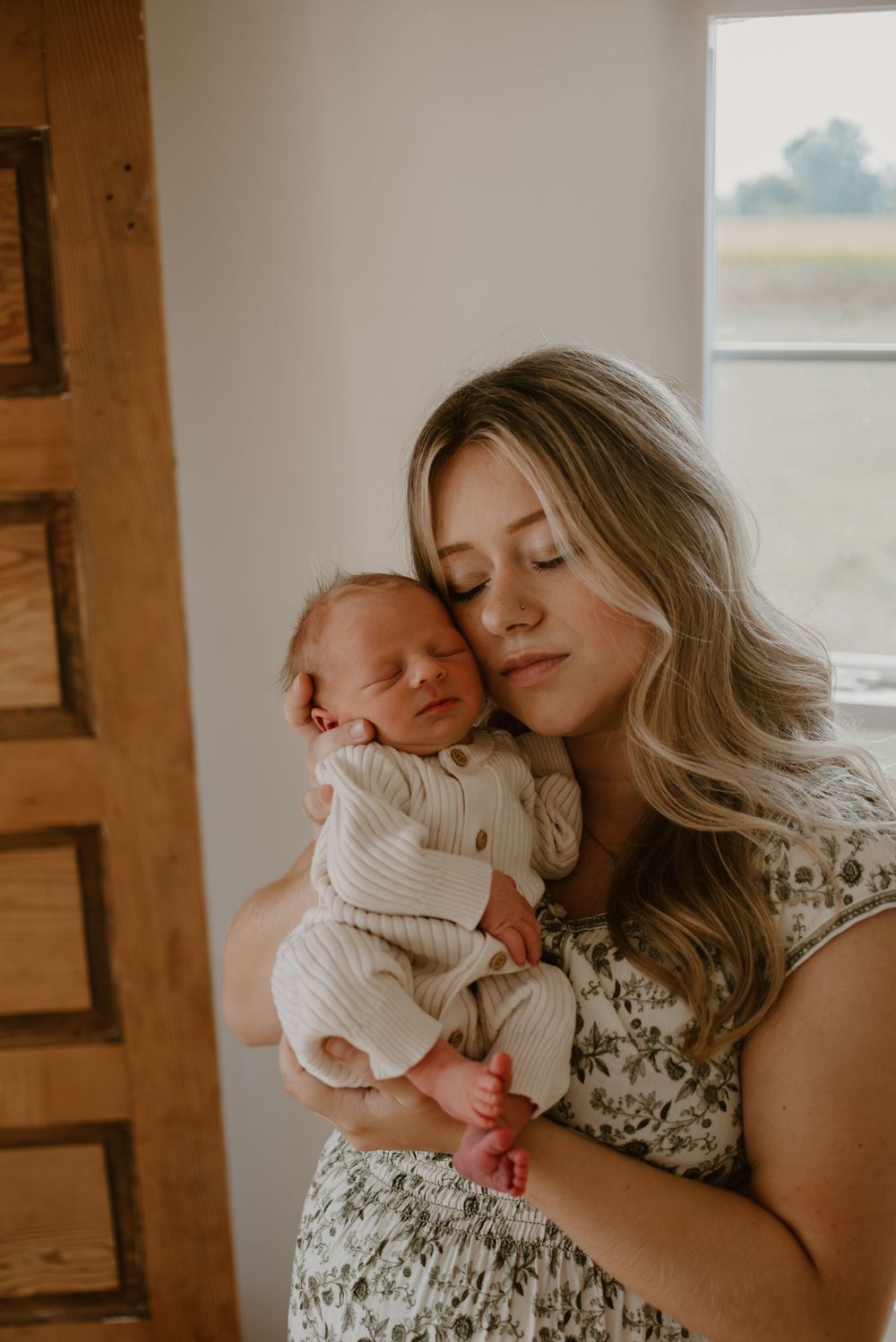 Tender moment between mother and newborn by window in warm natural light.