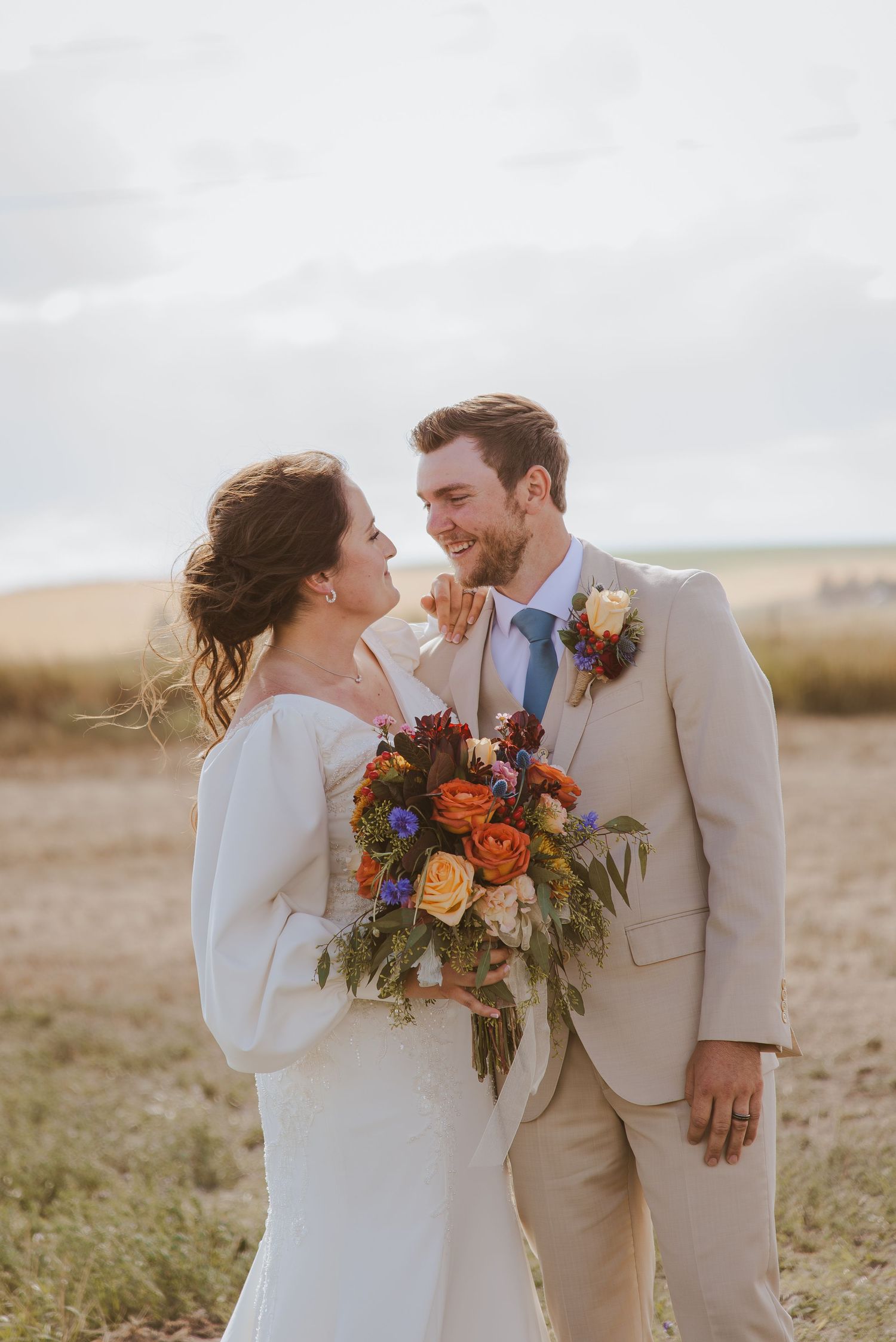 Newlyweds share a tender moment in a field at sunset with the bride holding a colorful fall bouquet.